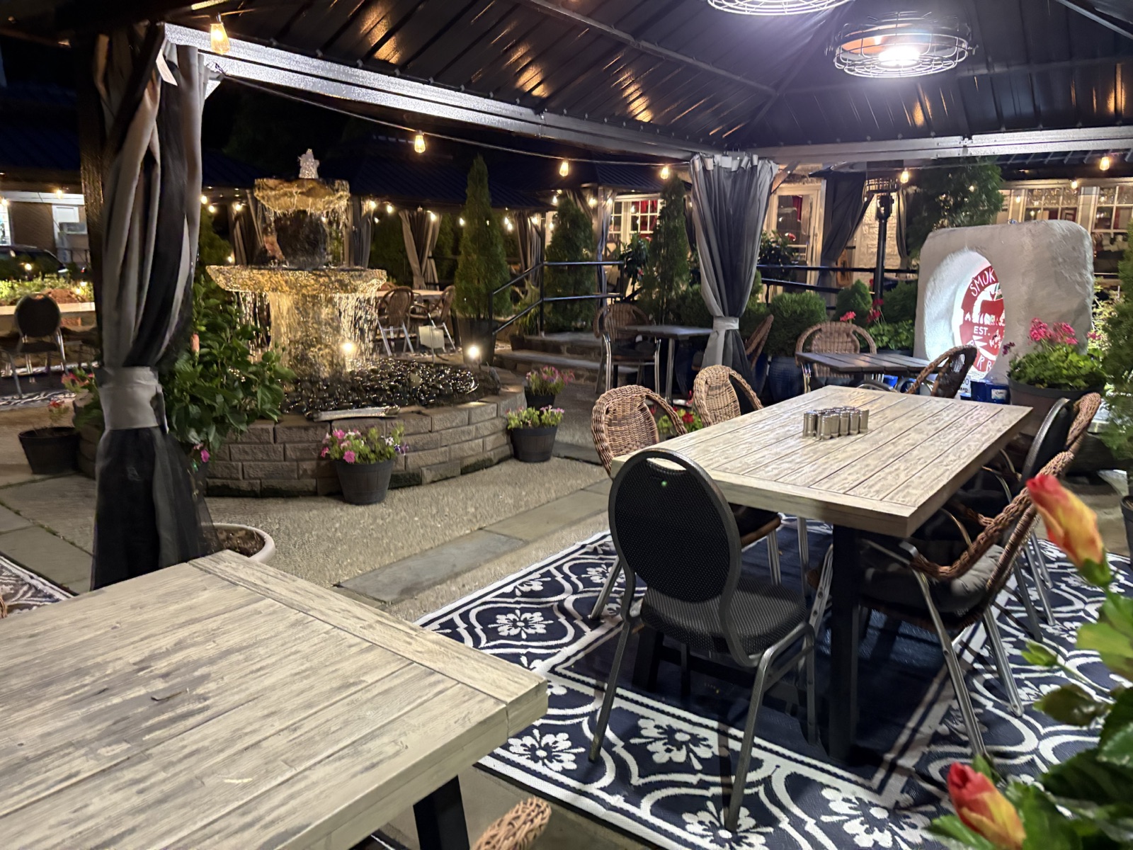Smoky Rock BBQ evening patio with a lit fountain, flowering planters, and covered outdoor tables